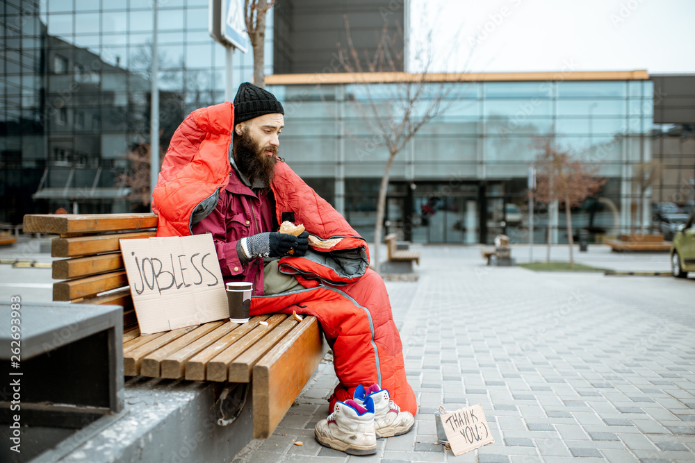 Depressed homeless beggar eating bread while sitting wrapped with sleeping bag on the bench near the