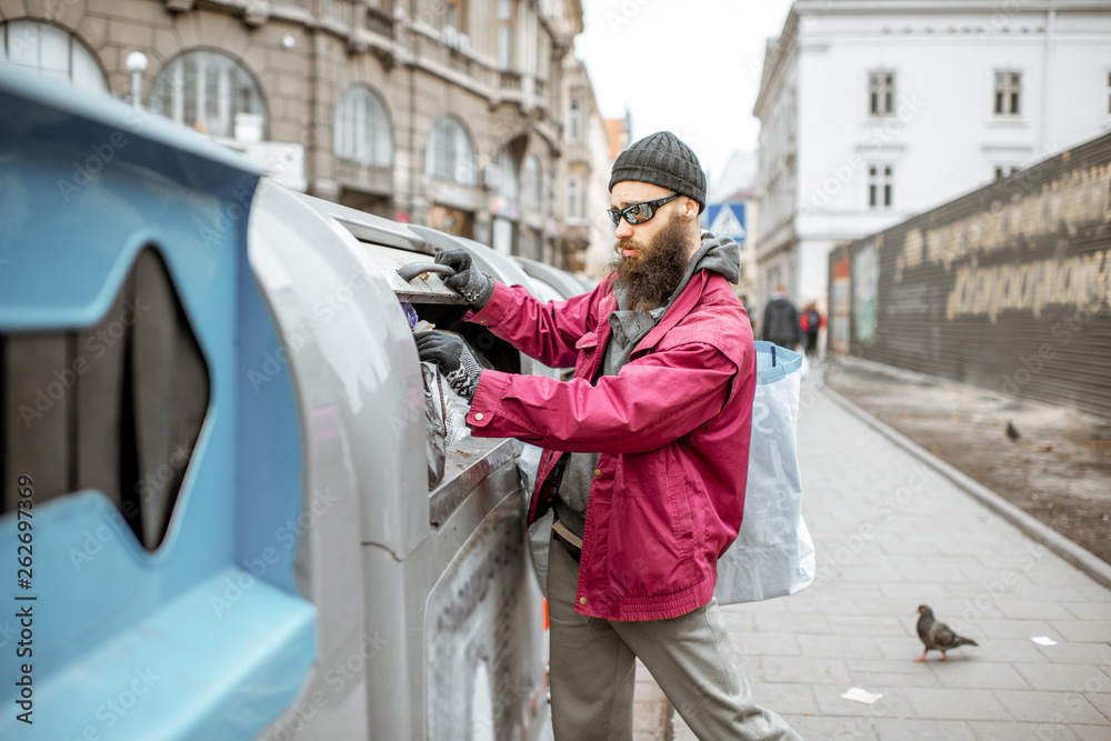 Homeless stylish beggar searching some food or valuable things in the trash containers in the old ci