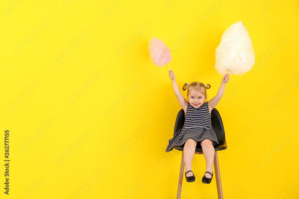 Cute little girl with cotton candy sitting on chair against color background