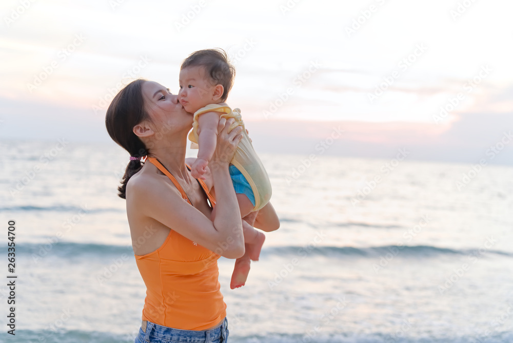 Asian mother standing on the beach holding her baby in two arms and raising the kid then kiss the ba