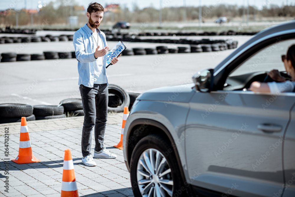 Male instructor teaching young woman driver to park a car on the training ground with traffic cones 