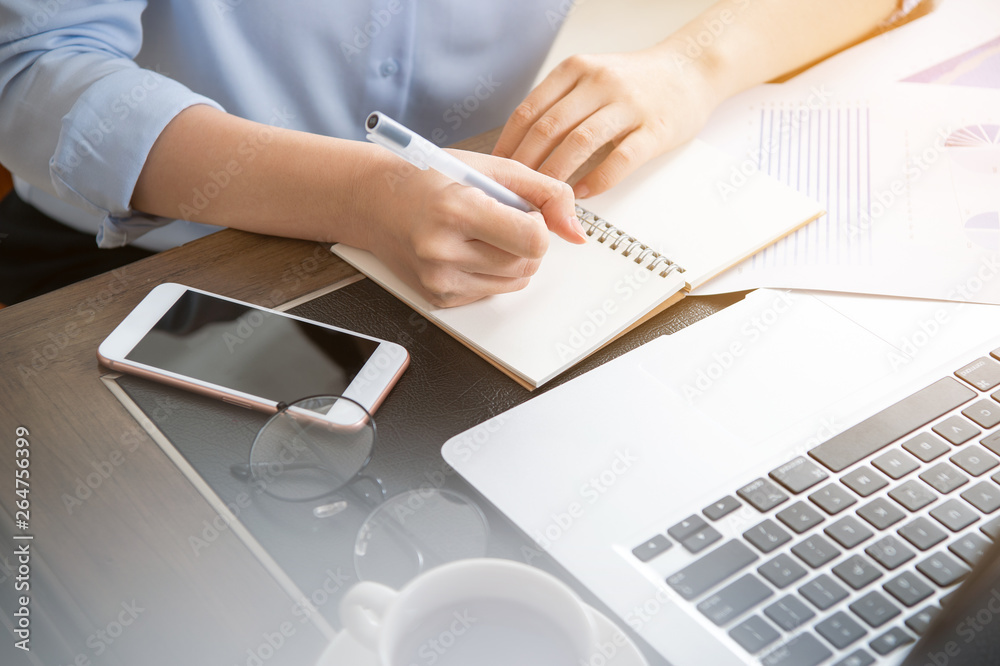 Business concept. Woman takes note for project with laptop and report in office desk. Backlighting, 