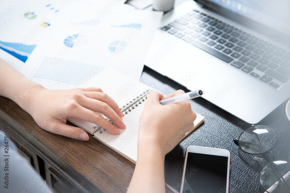 Business concept. Woman takes note for project with laptop and report in office desk. Backlighting, 