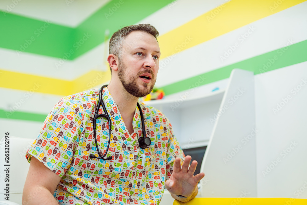 Doctor man in colorful shirt with stephoscope talking in his cabinet. Smart pediatrician carrying st