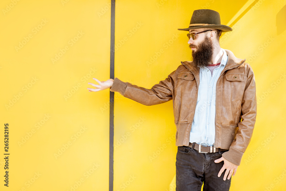 Portrait of a stylish bearded man dressed in jacket and hat on the bright yellow background outdoors