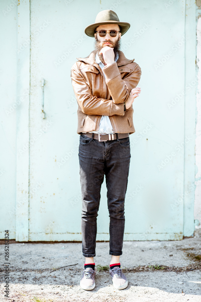 Full body portrait of a stylish bearded man dressed in jacket and hat on the light turquoise backgro