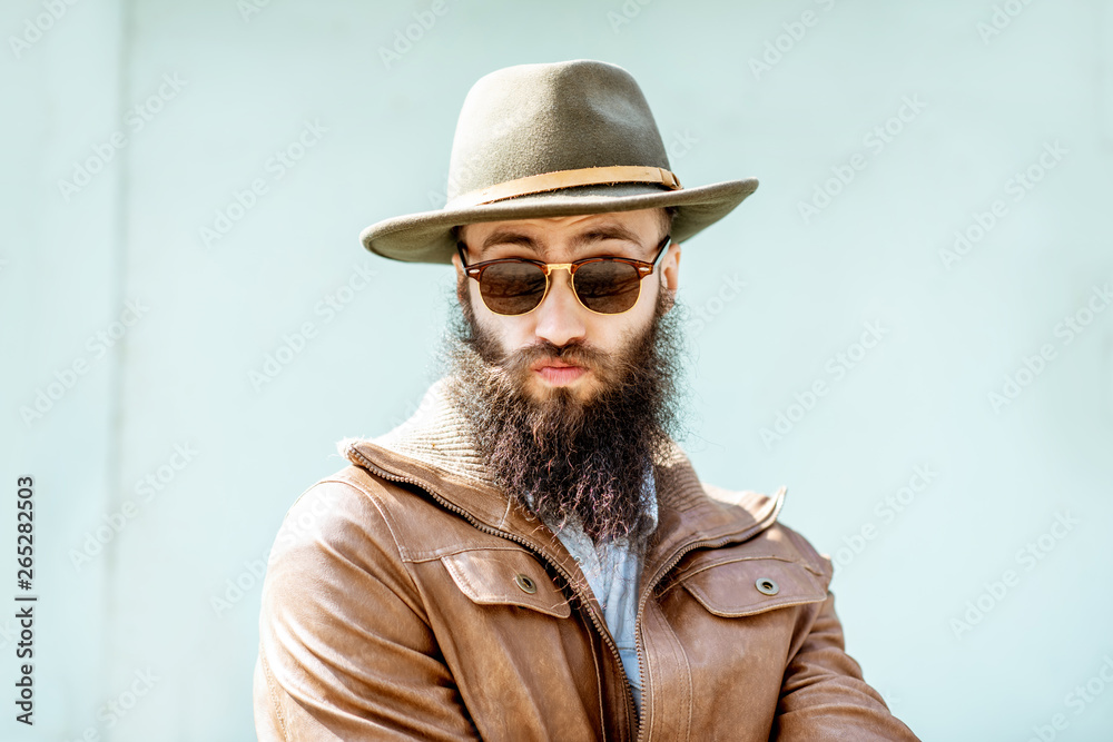 Portrait of a stylish bearded man dressed in jacket and hat on the light turquoise background outdoo