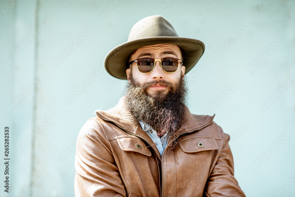 Portrait of a stylish bearded man dressed in jacket and hat on the light turquoise background outdoo