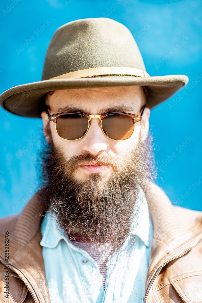 Close-up portrait of a stylish bearded man with hat and sunglasses on the blue wall background