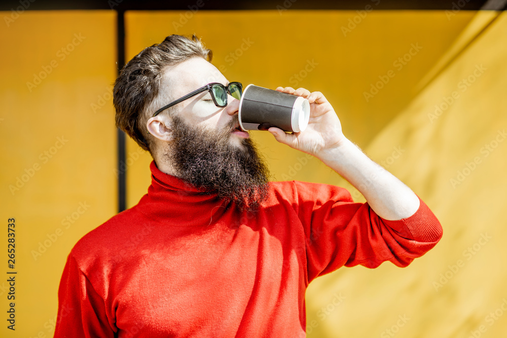 Portrait of a stylish bearded man in red swewater standing with coffee cup on the yellow background