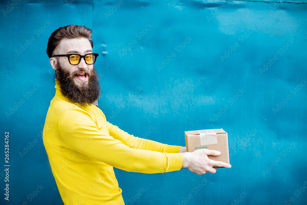 Stylish bearded man in yellow sweater standing with gift box on the blue background