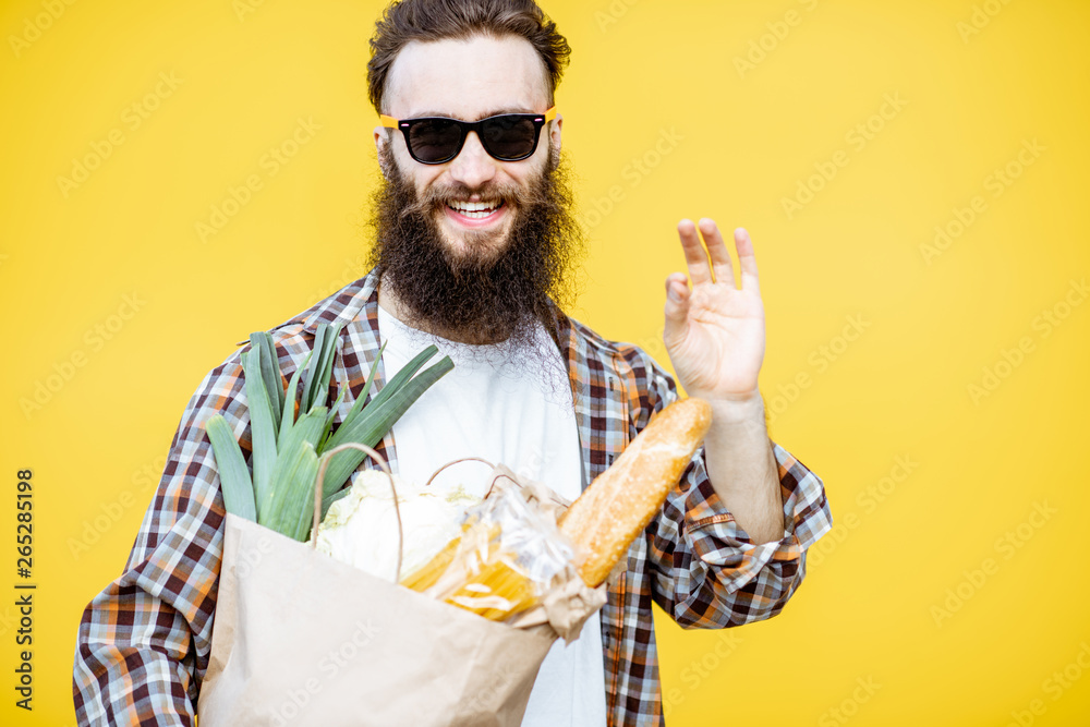 一个留着胡子的男人站在亮黄色的背上，手里拿着装满食物的购物纸袋的肖像