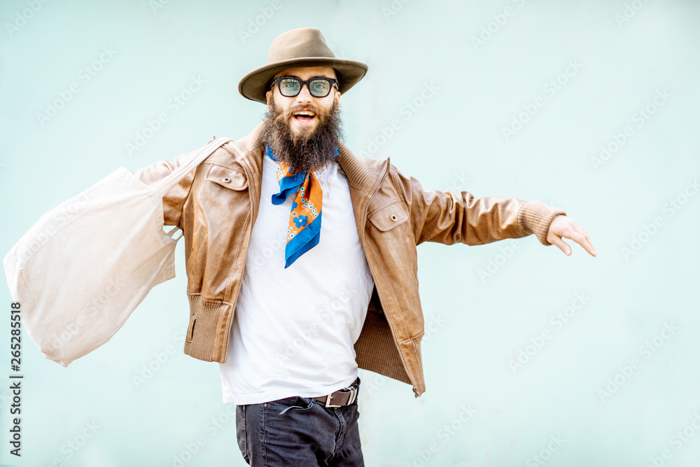 Portrait of a stylish man dressed in jacket, hat and scarf with bag on the turquoise background outd