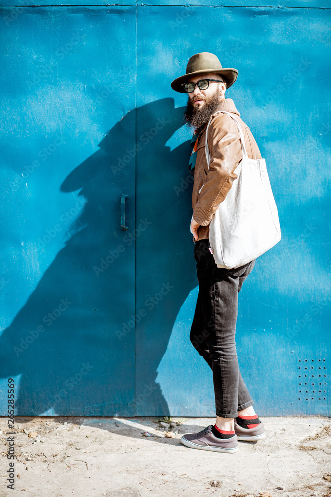 Portrait of a stylish bearded man dressed in jacket and hat standing with bag on the blue background