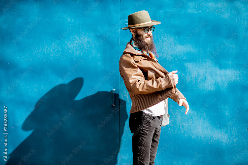 Portrait of a stylish bearded man dressed in jacket and hat standing with bag on the blue background