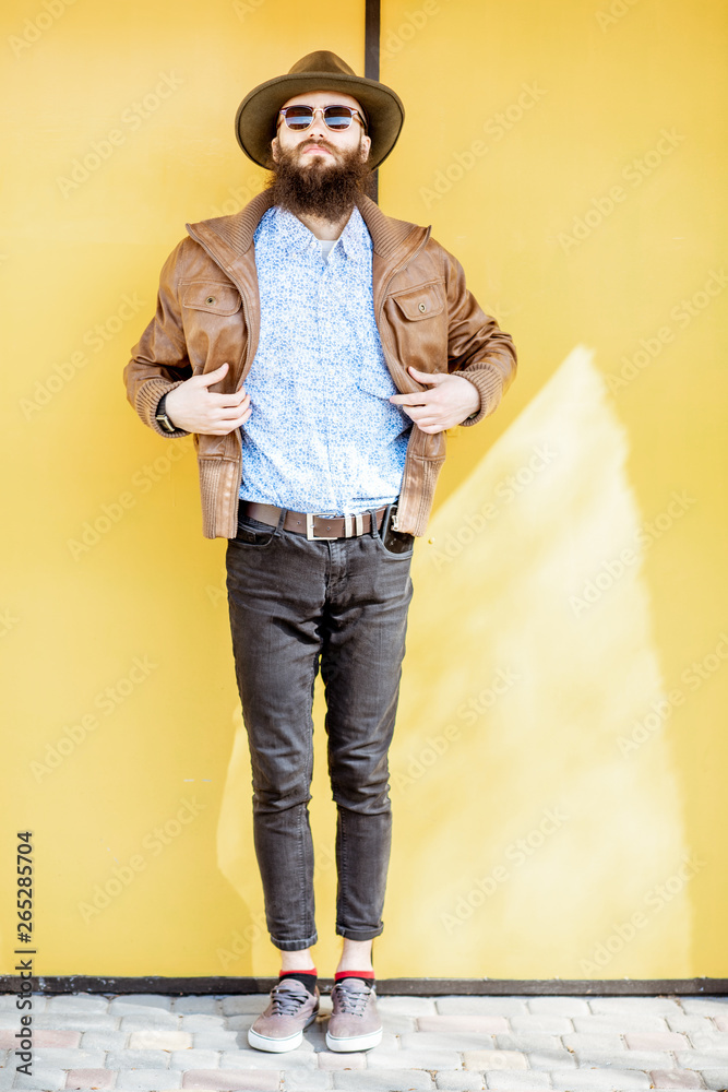 Full length portrait of a stylish bearded man dressed in jacket and hat on the bright yellow backgro