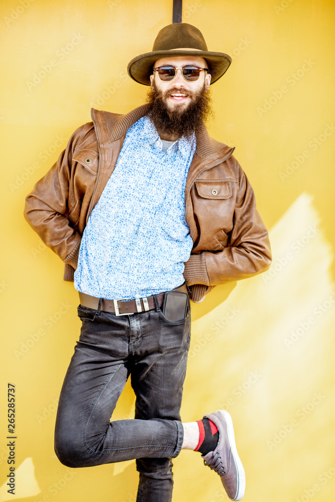 Full length portrait of a stylish bearded man dressed in jacket and hat on the bright yellow backgro