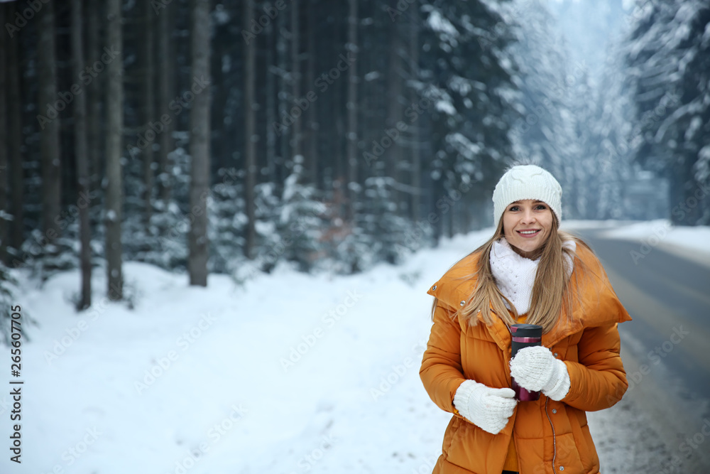 Beautiful young woman with hot drink near road in winter forest