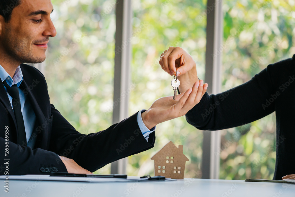 Adult agent giving key to smiling and happy business man while sitting with house model in office of