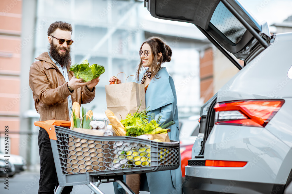 Young stylish couple with shopping cart full of fresh food, packing products into the car on the out