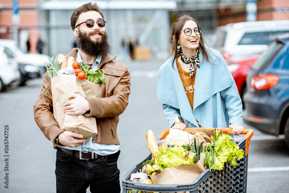 Young couple with shopping bags and cart full of fresh food on the outdooor parking near the superma