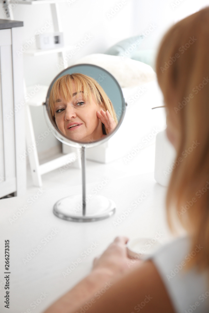 Mature woman with jar of cream looking in mirror at home