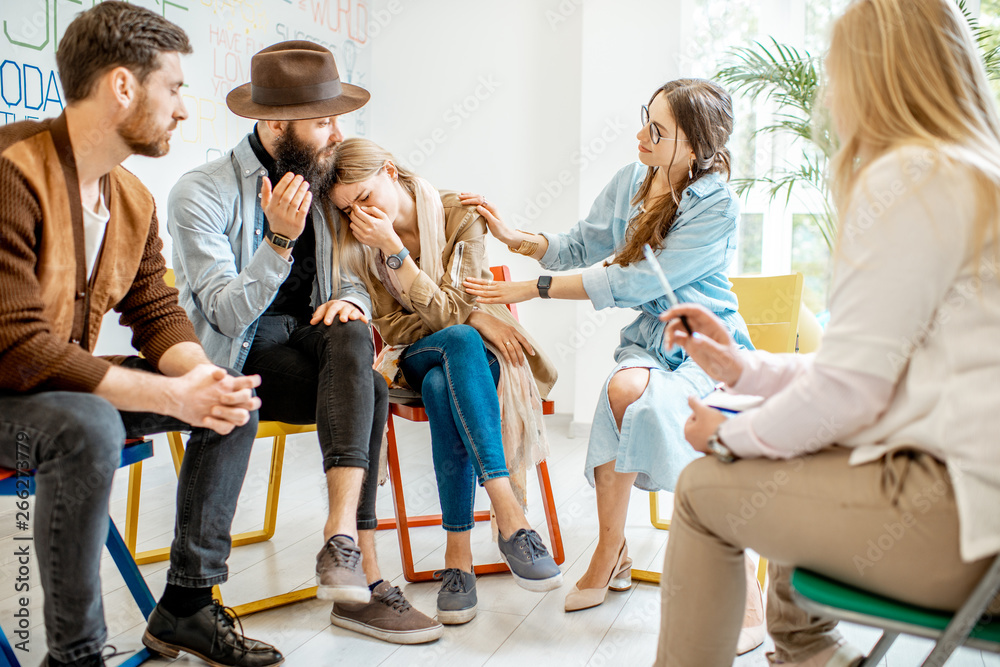 Young woman crying during the psychological therapy with group of people supporting her in the offic