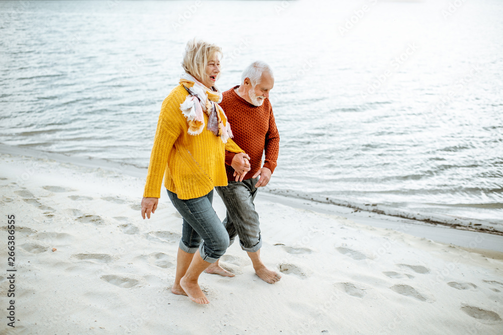 Lovely senior couple dressed in colorful sweaters walking on the sandy beach, enjoying free time dur