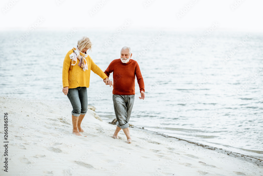 Lovely senior couple dressed in colorful sweaters walking on the sandy beach, enjoying free time dur