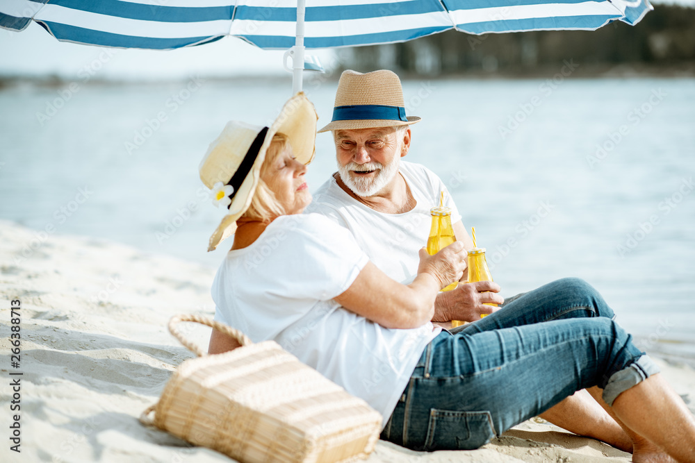 Happy senior couple relaxing, lying with drinks on the sandy beach, enjoying their retirement near t