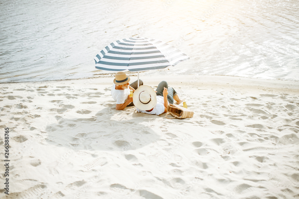 Senior couple sitting together under umbrella on the sandy beach, enjoying their retirement near the