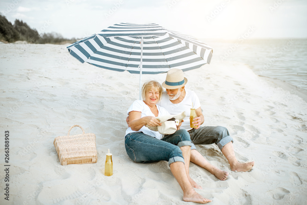 Happy senior couple relaxing, lying together under umbrella on the sandy beach, enjoying their retir