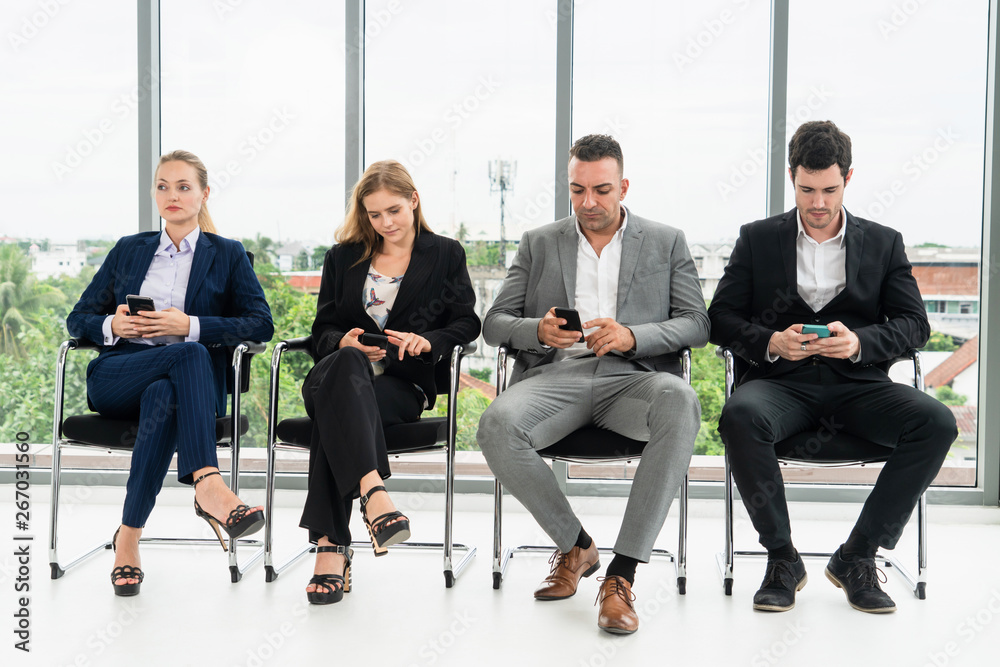 Businesswomen and businessmen using mobile phone while waiting on chairs in office for job interview