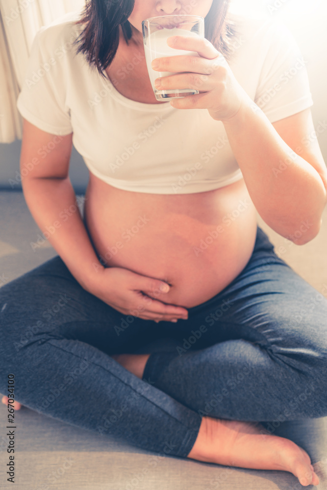 Happy pregnant woman drinks milk in glass at home while taking care of her child. The young expectin