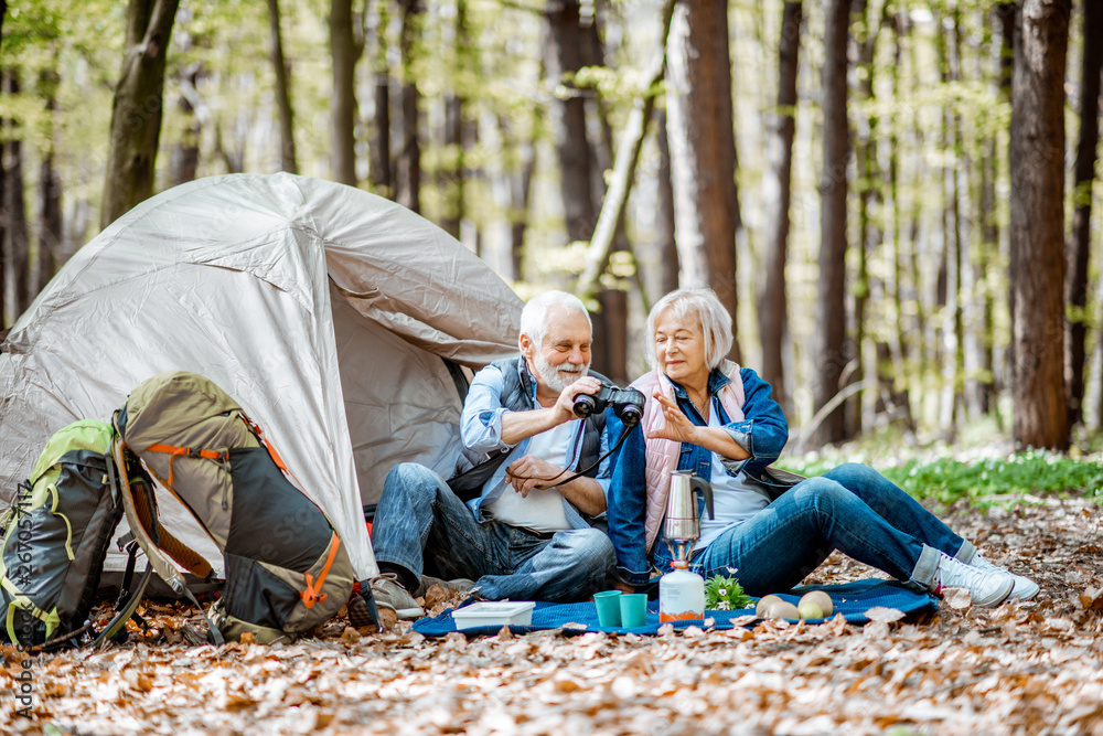 Senior couple sitting together at the campsite with tent and backpacks, enjoying nature with binocul