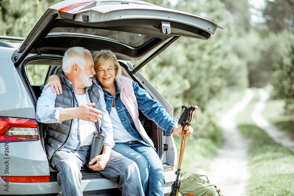 Senior couple sitting at the car trunk, enjoying nature while traveling in the young pine forest