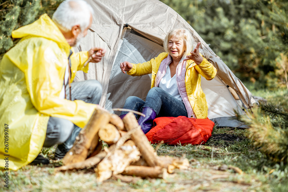 Senior couple in yellow raincoats having fun while making fireplace at the campsite near the tent in