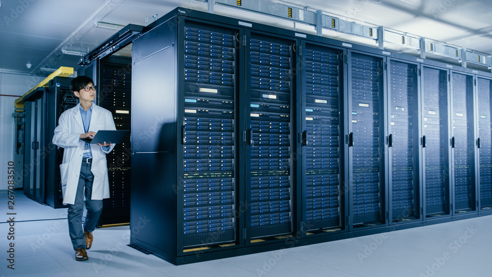 In Data Center: Male IT Specialist Wearing White Coat Walks Alongside Row of Server Racks, Uses Lapt