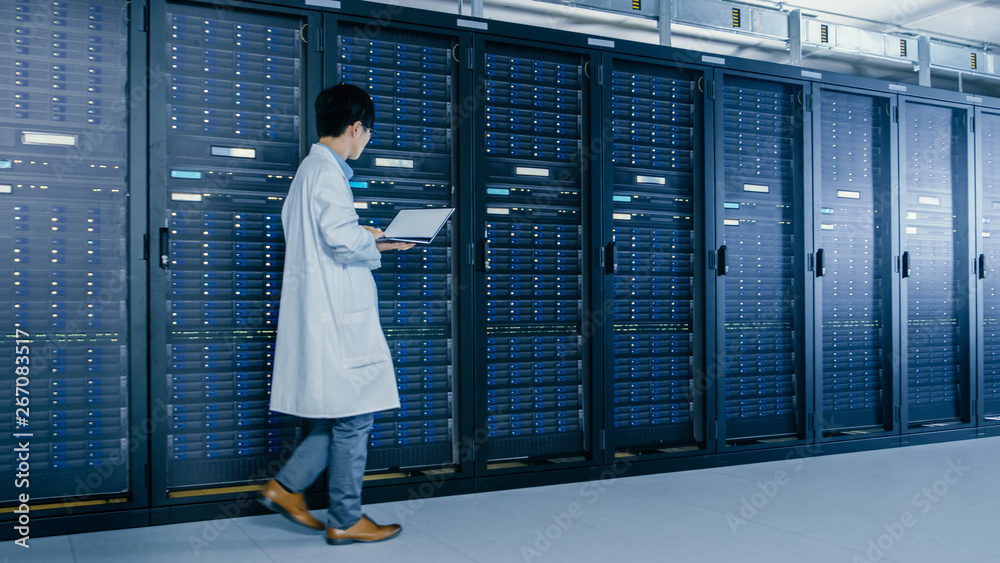 In Data Center: Male IT Specialist Wearing White Coat Walks Alongside Row of Server Racks, Uses Lapt