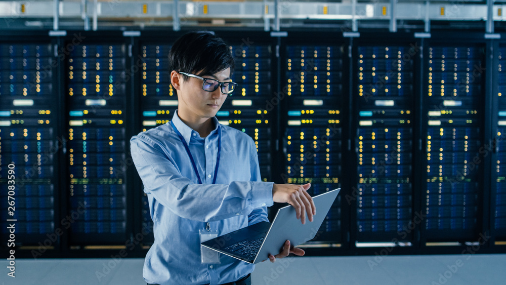 In Dark Data Center: Male IT Specialist Stands Beside Row of Operational Server Racks, Uses Laptop f