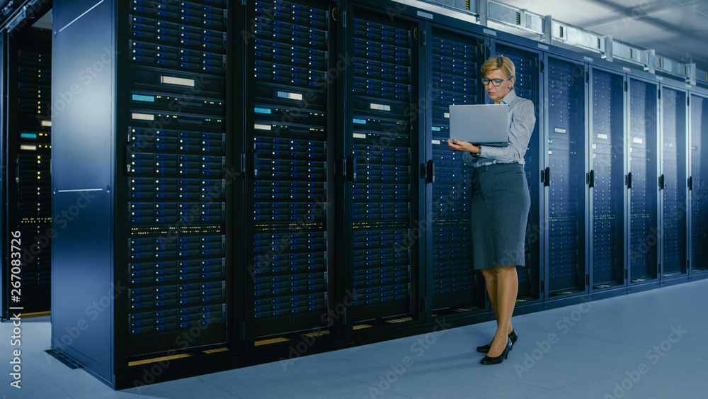 In Data Center: Female IT Technician Stands Near Rack Cabinet and Runs Maintenance Program on Laptop