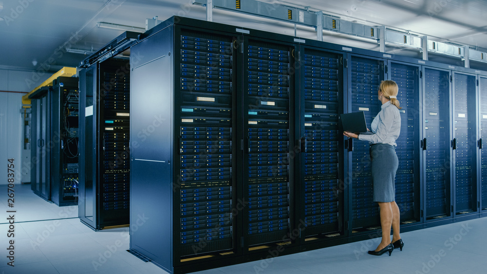 In Data Center: Female IT Technician Stands Near Rack Cabinet and Runs Maintenance Program on Laptop