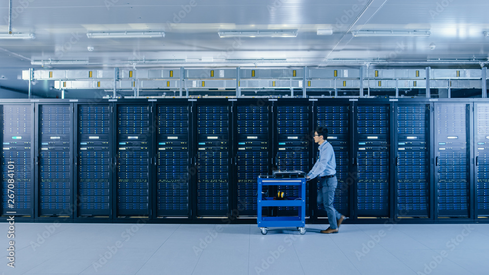 In the Modern Data Center: IT Technician Working with Server Racks, Pushes Cart Between Rows of Serv