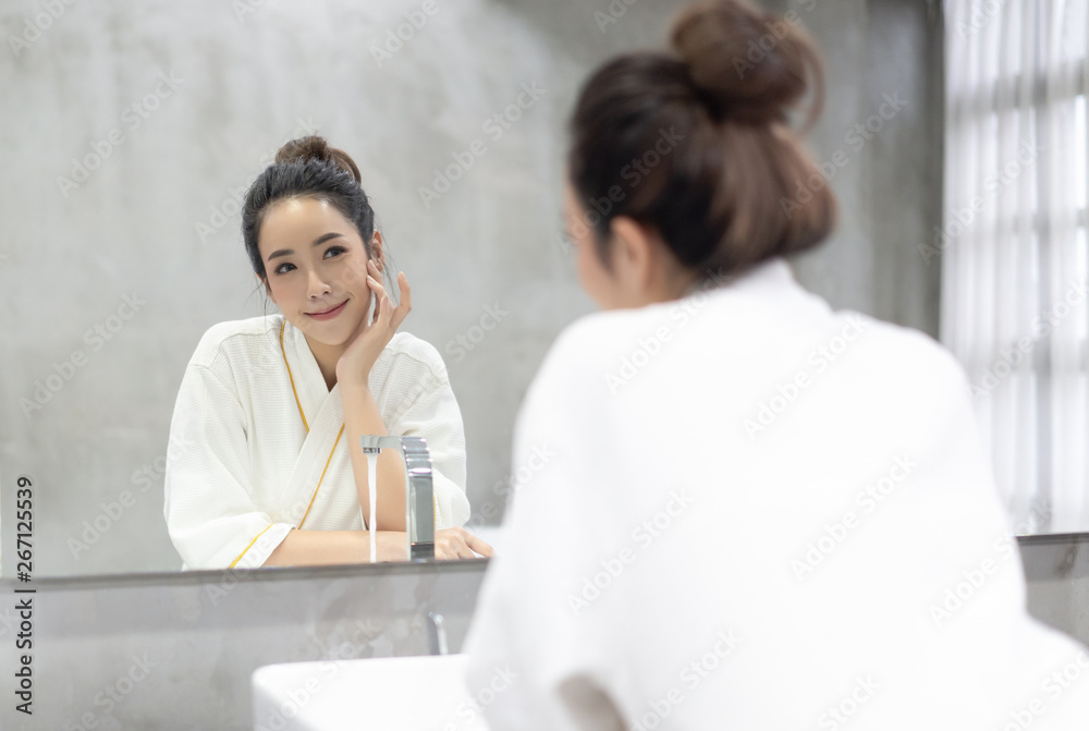 Facial Beauty.Beautiful smiling young Asian woman in bathrobe applying moisturizer cream on her pret