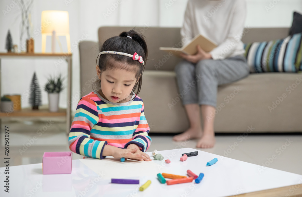 Little child girl playing with a colorful toys modeling clay on the floor, at background mom is sitt