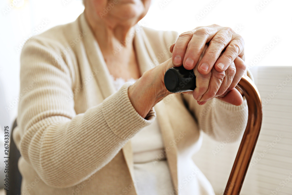 Elderly woman sitting in nursing home room holding walking quad cane with wrinked hand. Old age seni