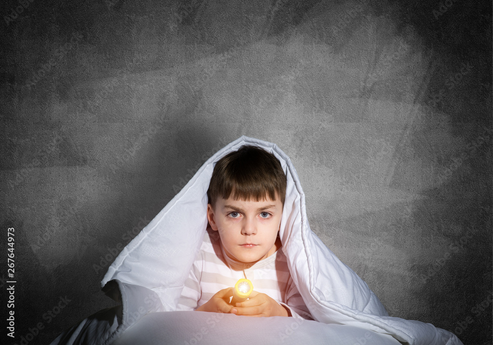 Serious child with flashlight hiding under blanket