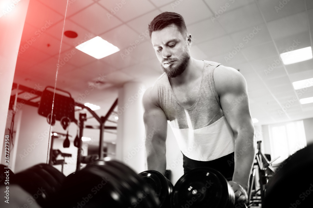 Black and white photo of a strong bodybuilder standing at the dumbbells in gym. Handsome man with mu