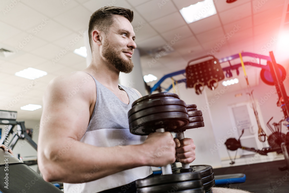 Handsome smiling man sitting with two heavy dumbbells in the crossfit gym. Strong bodybuilder is hol