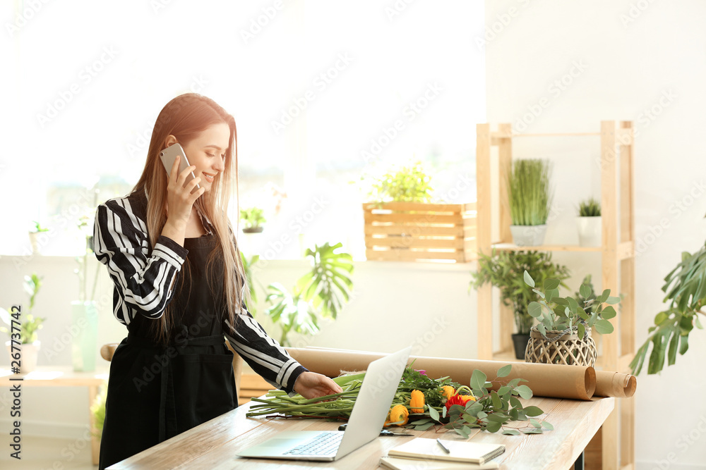 Young florist talking by phone while working with laptop in shop